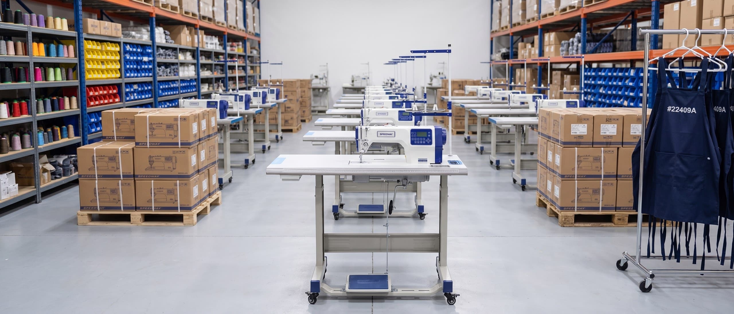 Speedway distribution warehouse — a SPEEDWAY AI4 lockstitch machine on its table in the foreground, rows of new machines and boxed inventory on pallets behind, navy work aprons on a rack