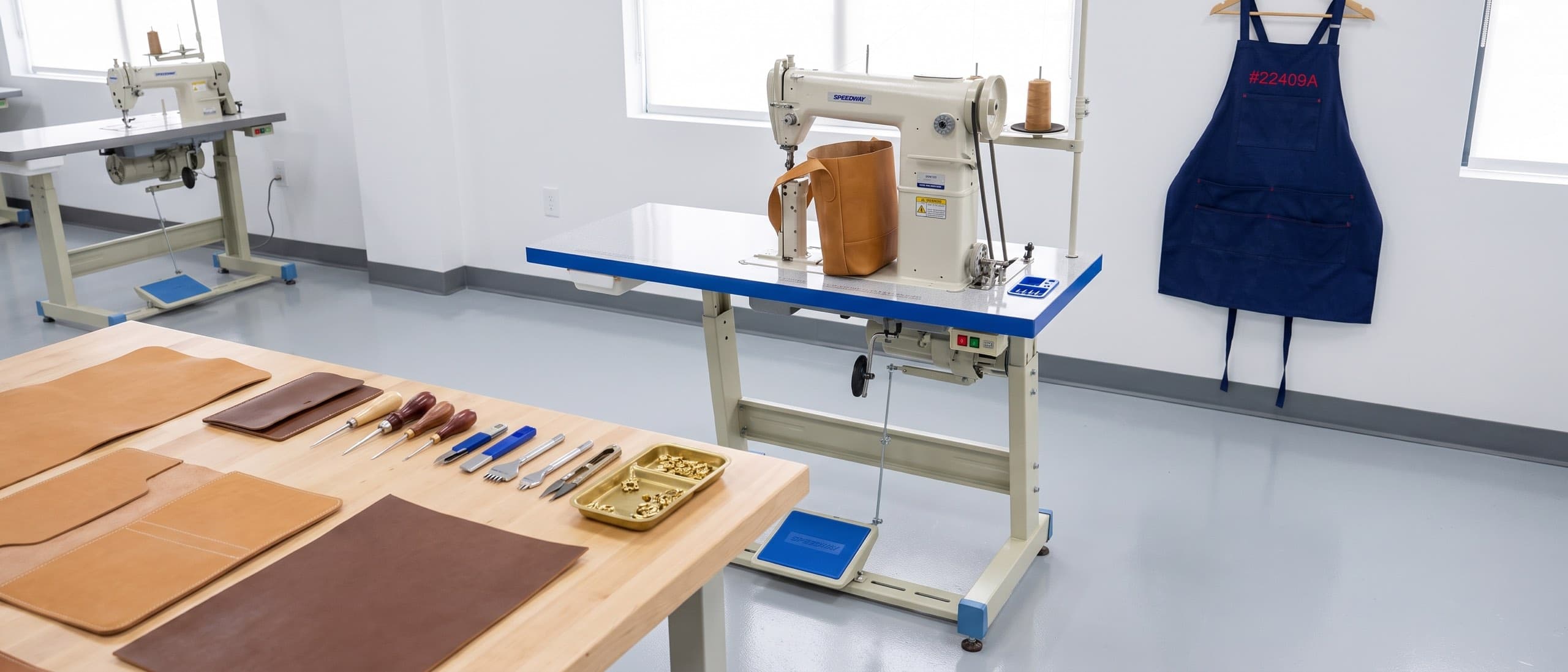 Modern leather goods workshop — SPEEDWAY SW-810 post-bed machine with a tan leather bag at the post, cut panels and edge tools on the cutting table